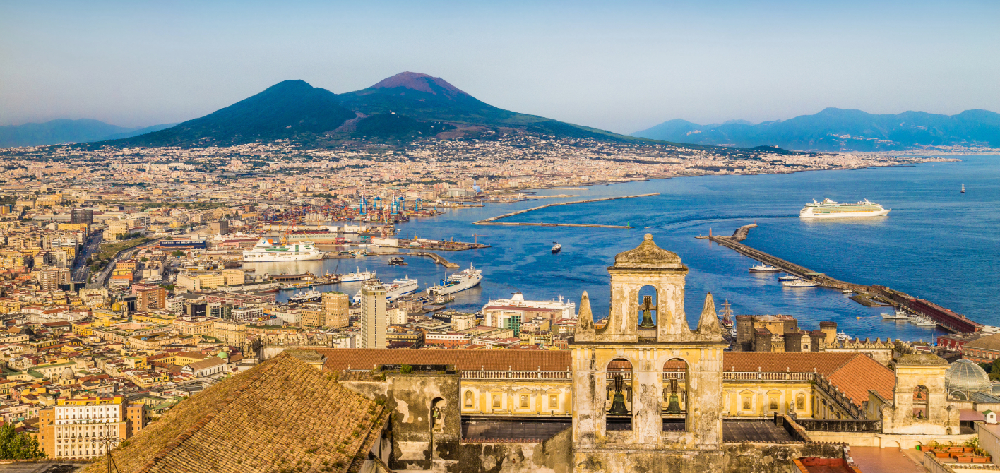 Luftaufnahme von Neapel, Italien, mit dem markanten Vesuv im Hintergrund. Im Vordergrund befindet sich das Dach einer historischen Kirche, dahinter erstrecken sich die Stadt, der Hafen und der tiefblaue Golf von Neapel.
