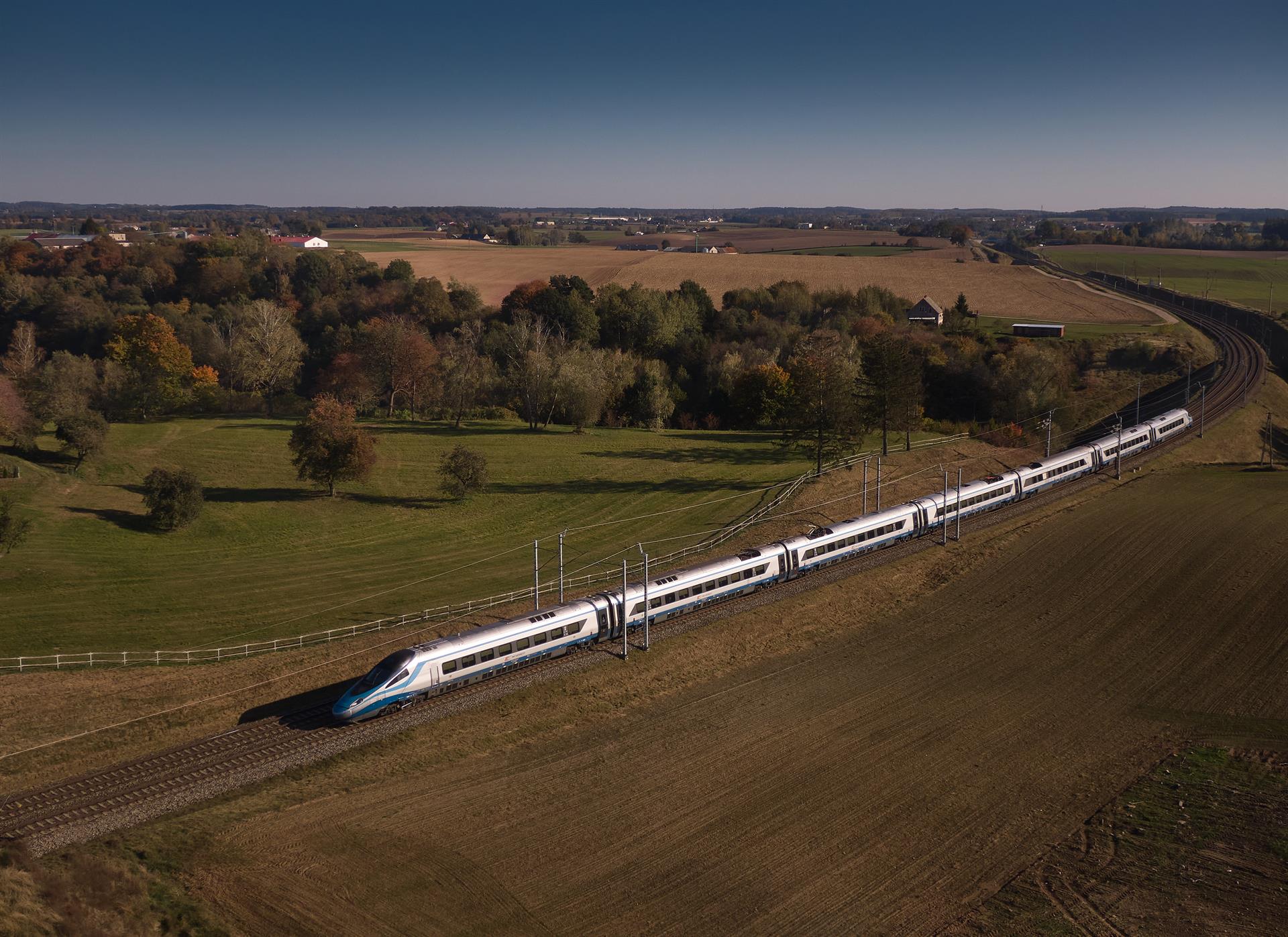 Ein weiß-blauer Pendolino-Hochgeschwindigkeitszug fährt aus der Vogelperspektive durch eine herbstliche, ländliche Landschaft mit Feldern und bunten Laubbäumen. 