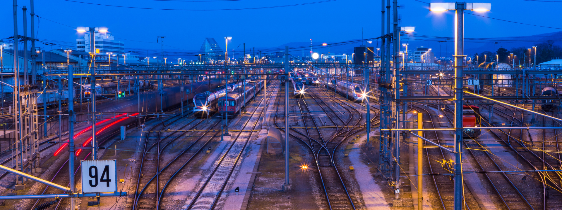 Weitläufiges SBB-Gleisfeld bei Nacht mit zahlreichen beleuchteten Oberleitungsmasten, parkenden Regionalzügen und roten Lichtspuren eines fahrenden Zuges, das die Komplexität des Schweizer Schienennetzes zeigt.