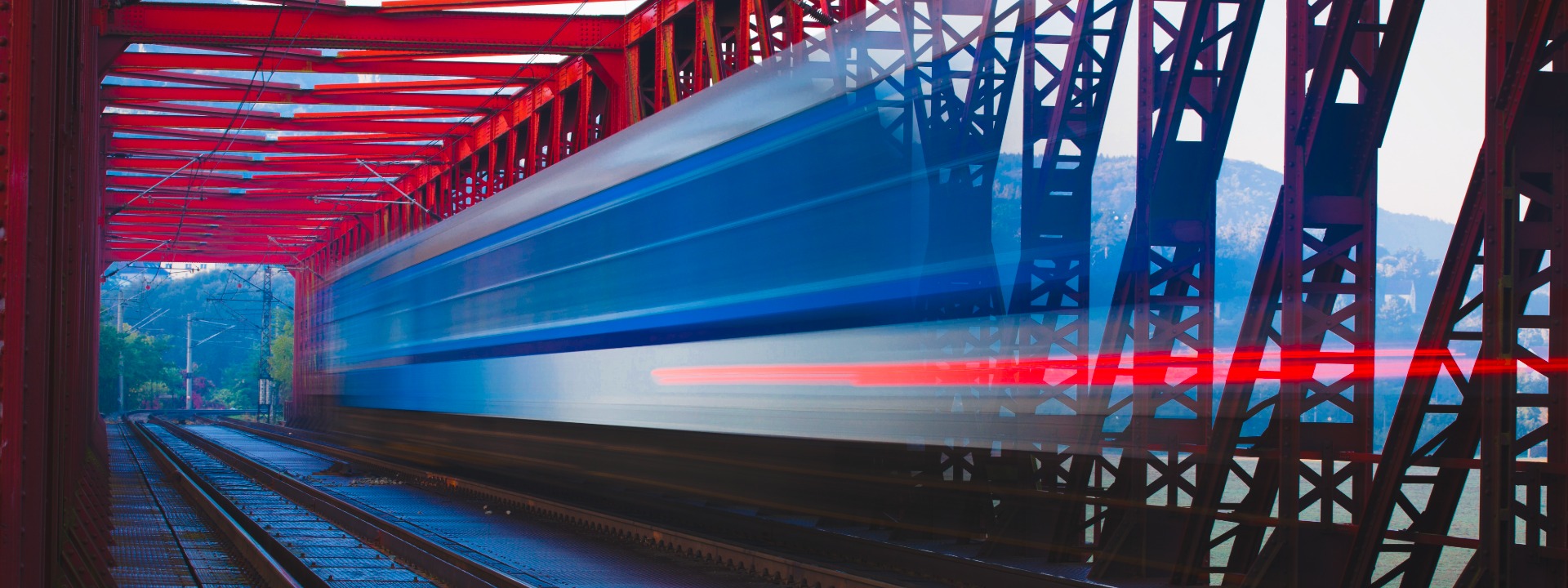 Ein schneller, blau-weißer Pendolino-Zug in Bewegungsunschärfe auf einer leuchtend roten Stahlgitterbrücke, die durch eine grüne, hügelige Landschaft führt.