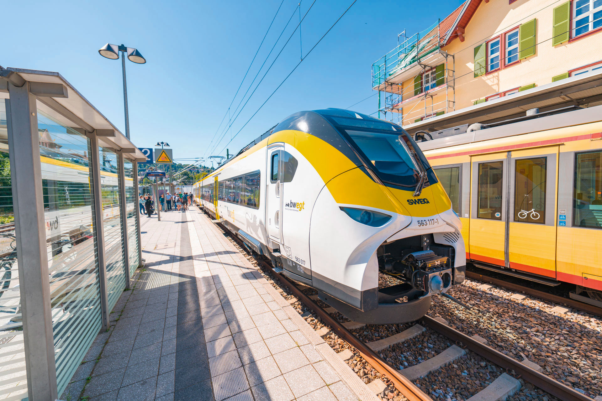 Ein moderner, weiß-gelber Triebzug vom Typ Siemens Mireo steht bei Sonnenschein an einem Bahnsteig, während Fahrgäste im Hintergrund warten