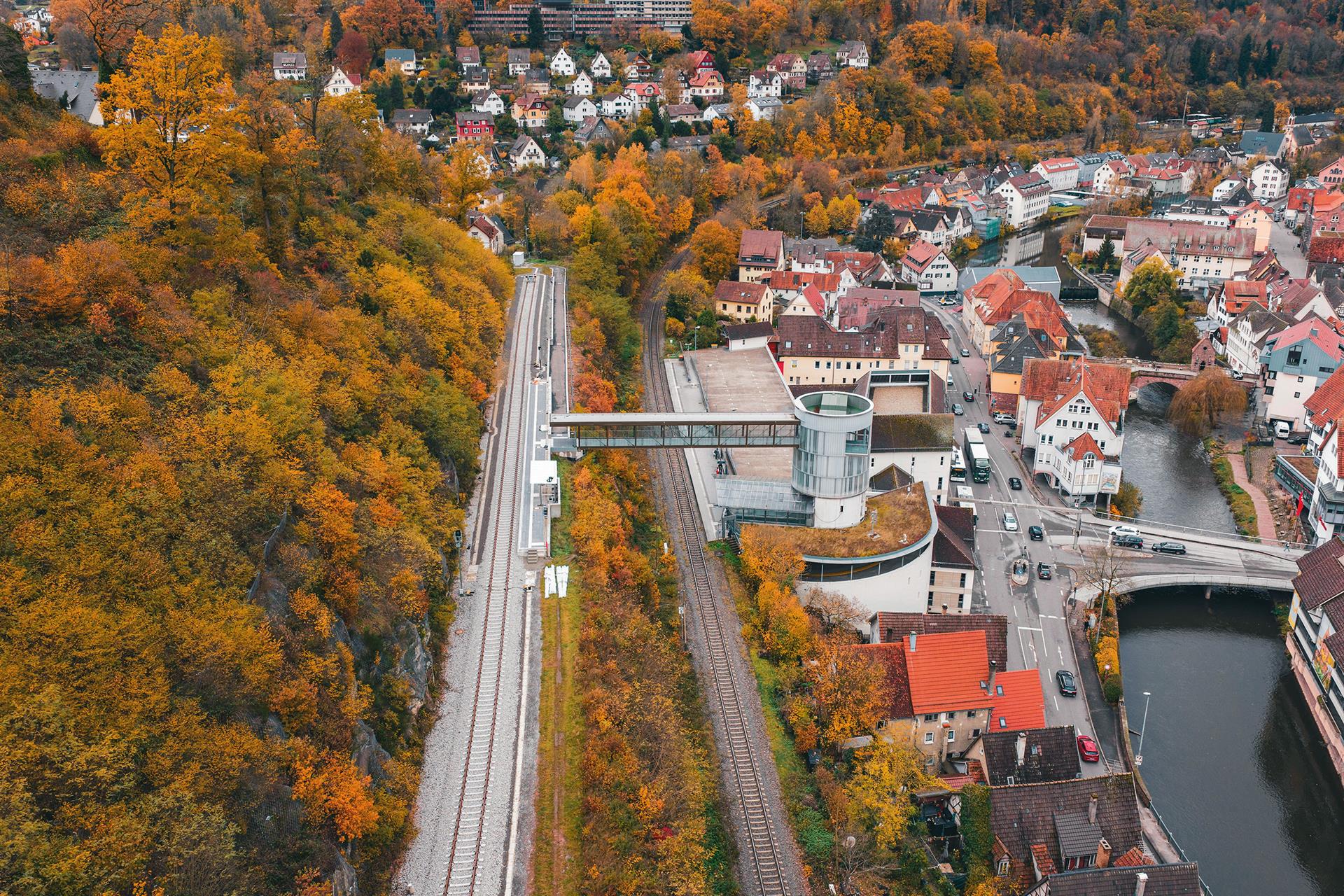 Luftaufnahme des Calwer Bahnhofs im Herbst: Neben den Gleisen im Tal führt eine Stahl-Glas-Brücke von einem Parkhaus zu einem neu gebauten, höher gelegenen Gleisbett am bewaldeten Hang