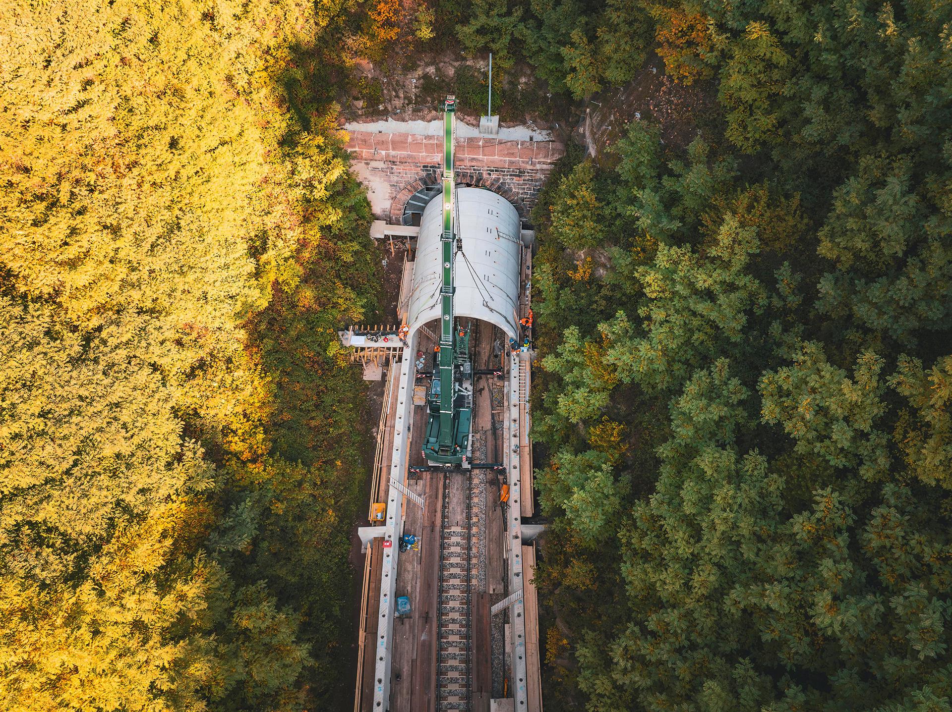 Luftaufnahme einer Gleisbaustelle im herbstlichen Wald: Ein grüner Schienenkran hebt ein großes, gewölbtes Betonelement vor ein Tunnelportal der Hermann-Hesse-Bahn, um eine Einhausung für den Fledermausschutz zu errichten