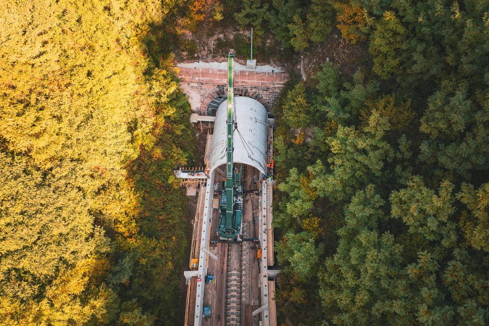 Luftaufnahme einer Gleisbaustelle im herbstlichen Wald: Ein grüner Schienenkran hebt ein großes, gewölbtes Betonelement vor ein Tunnelportal der Hermann-Hesse-Bahn, um eine Einhausung für den Fledermausschutz zu errichten
