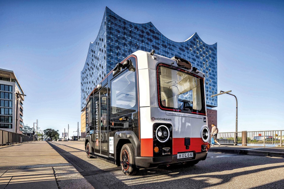 Ein futuristischer, weiß-rot-schwarzer Holon Shuttle-Bus der Hamburger Hochbahn steht bei sonnigem Wetter auf einer Straße direkt vor der modernen, wellenförmigen Glasfassade der Elbphilharmonie Hamburg.