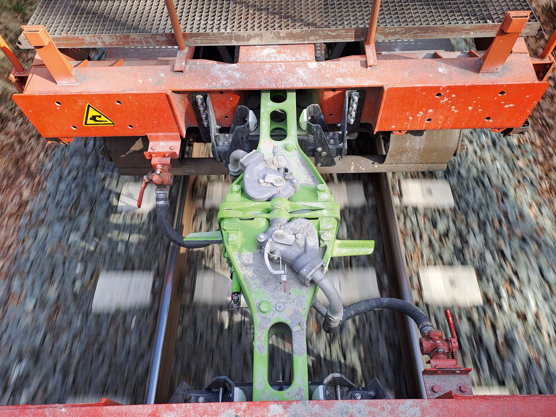 Nahaufnahme einer grünen Digitalen Automatischen Kupplung zwischen zwei roten Güterwagen während der Fahrt, mit unscharfem Gleisbett im Hintergrund.