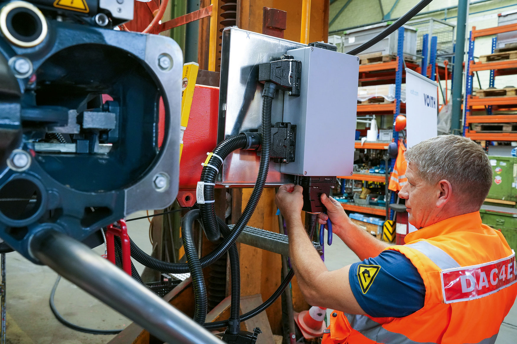 Ein Techniker mit orangefarbener Warnweste und der Aufschrift DAC4EU arbeitet in einer Werkstatt an der Verkabelung eines grauen Schaltkastens an einer Maschinenkomponente.