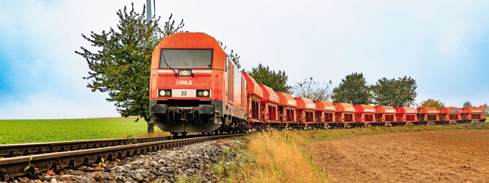 Rote WLE-Diesellokomotive Eurorunner zieht beladene Schüttgutwagen auf einer eingleisigen Bahnstrecke durch eine grüne Landschaft mit Windrad im Hintergrund.