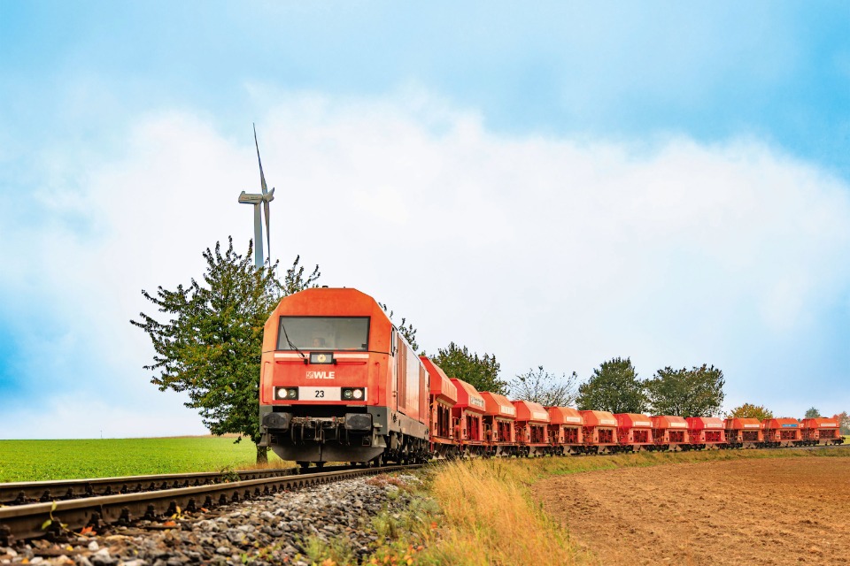 Rote WLE-Diesellokomotive Eurorunner zieht beladene Schüttgutwagen auf einer eingleisigen Bahnstrecke durch eine grüne Landschaft mit Windrad im Hintergrund.