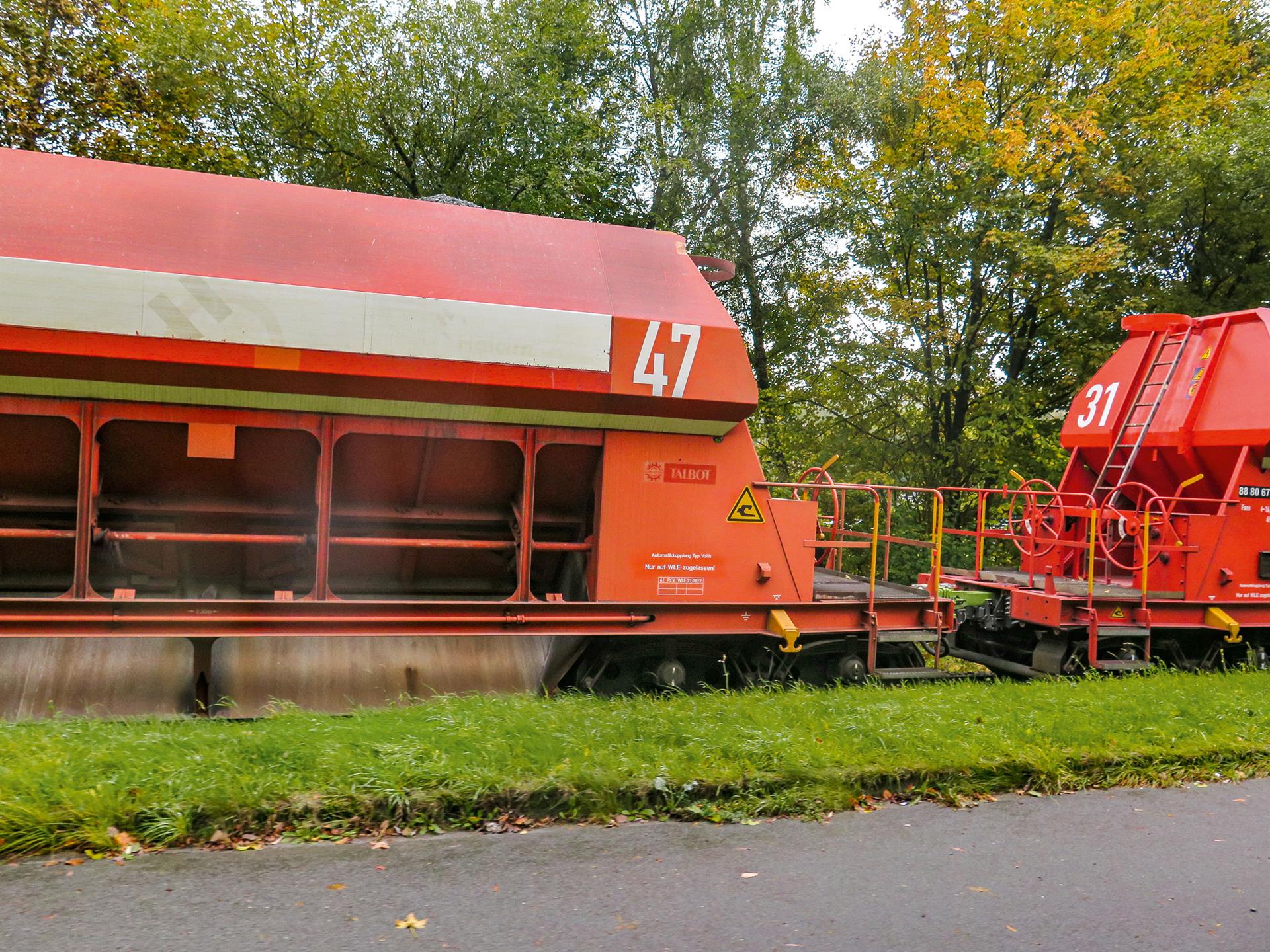 Ein WLE-Mitarbeiter in oranger Warnkleidung und Helm steht auf der Plattform eines roten Schüttgutwagens mit grüner Digitaler Automatischer Kupplung in einer staubigen Verladeanlage.