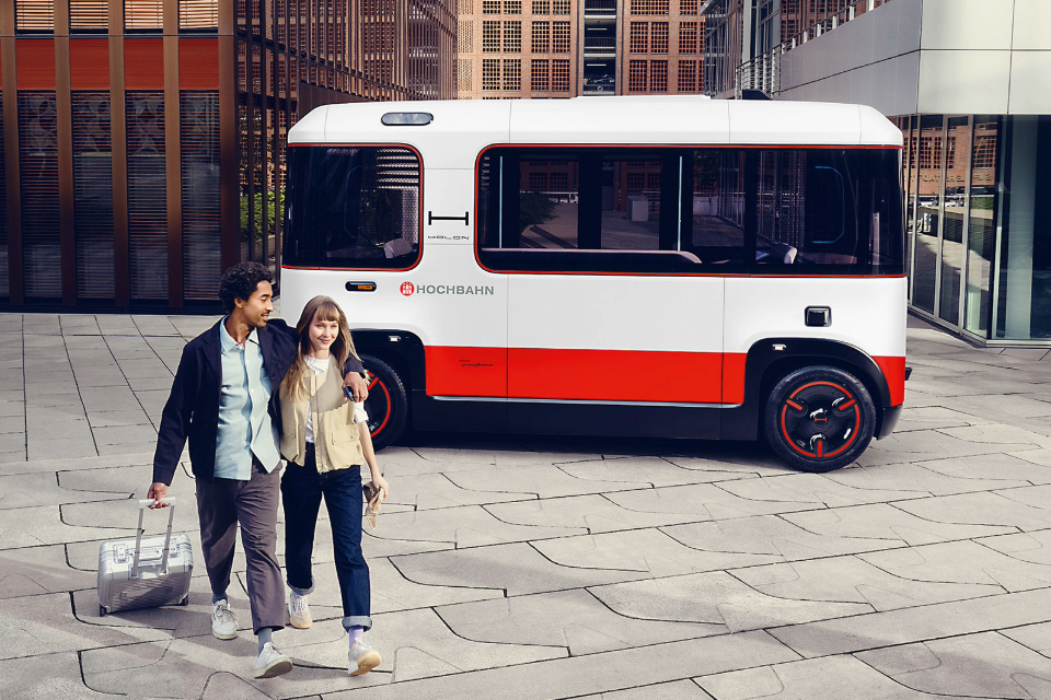 Ein Paar spaziert durch eine moderne Stadt, daneben parkt ein autonomer Shuttle-Bus der Hamburger Hochbahn, Modell Holon, vor einer urbanen Kulisse mit Bürogebäuden.