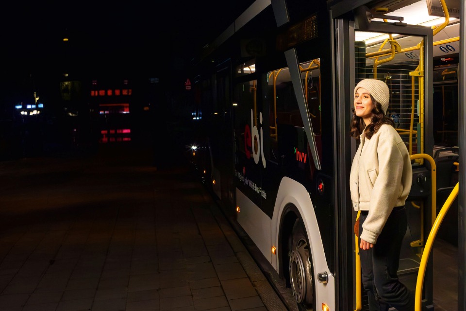 Eine junge Frau mit heller Jacke und Mütze steht nachts in der geöffneten Tür eines hvv-Linienbusses, bereit zum Aussteigen an einem dunklen Straßenrand.