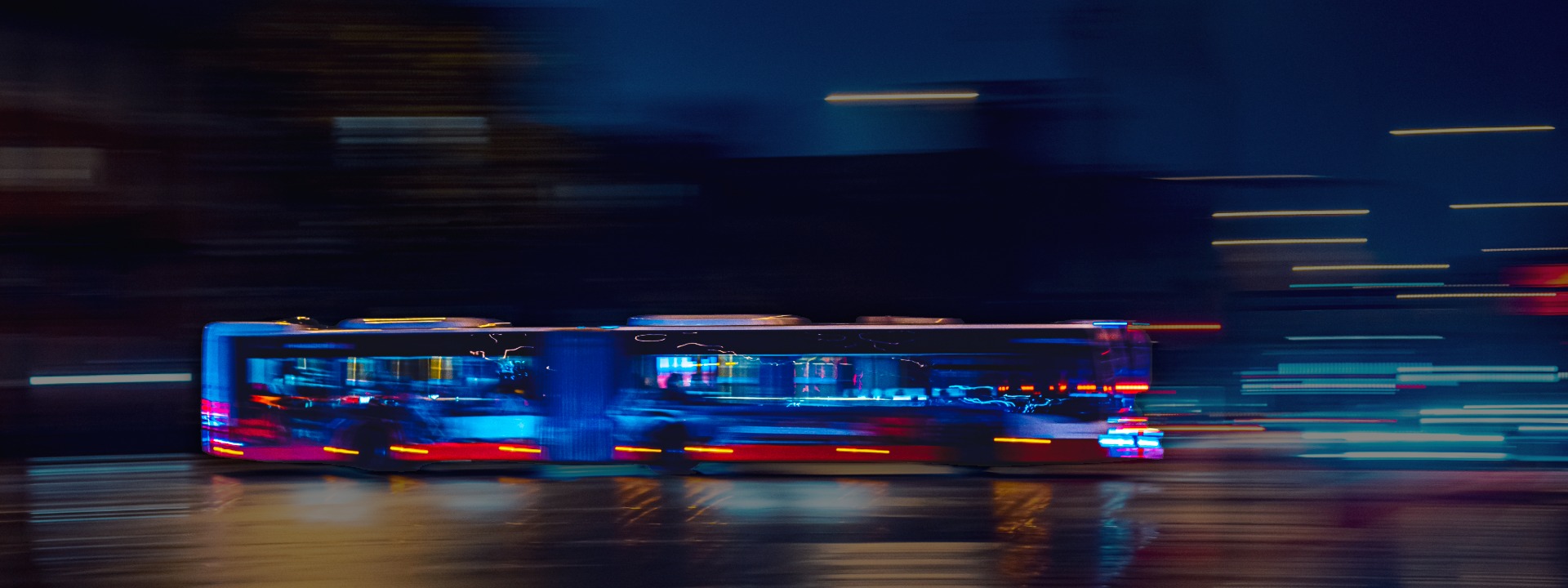 Bewegungsunscharfe Aufnahme eines Linienbusses bei Nacht in einer Stadt. Die Lichter des Busses und der Umgebung ziehen blaue und rote Streifen und vermitteln Geschwindigkeit.