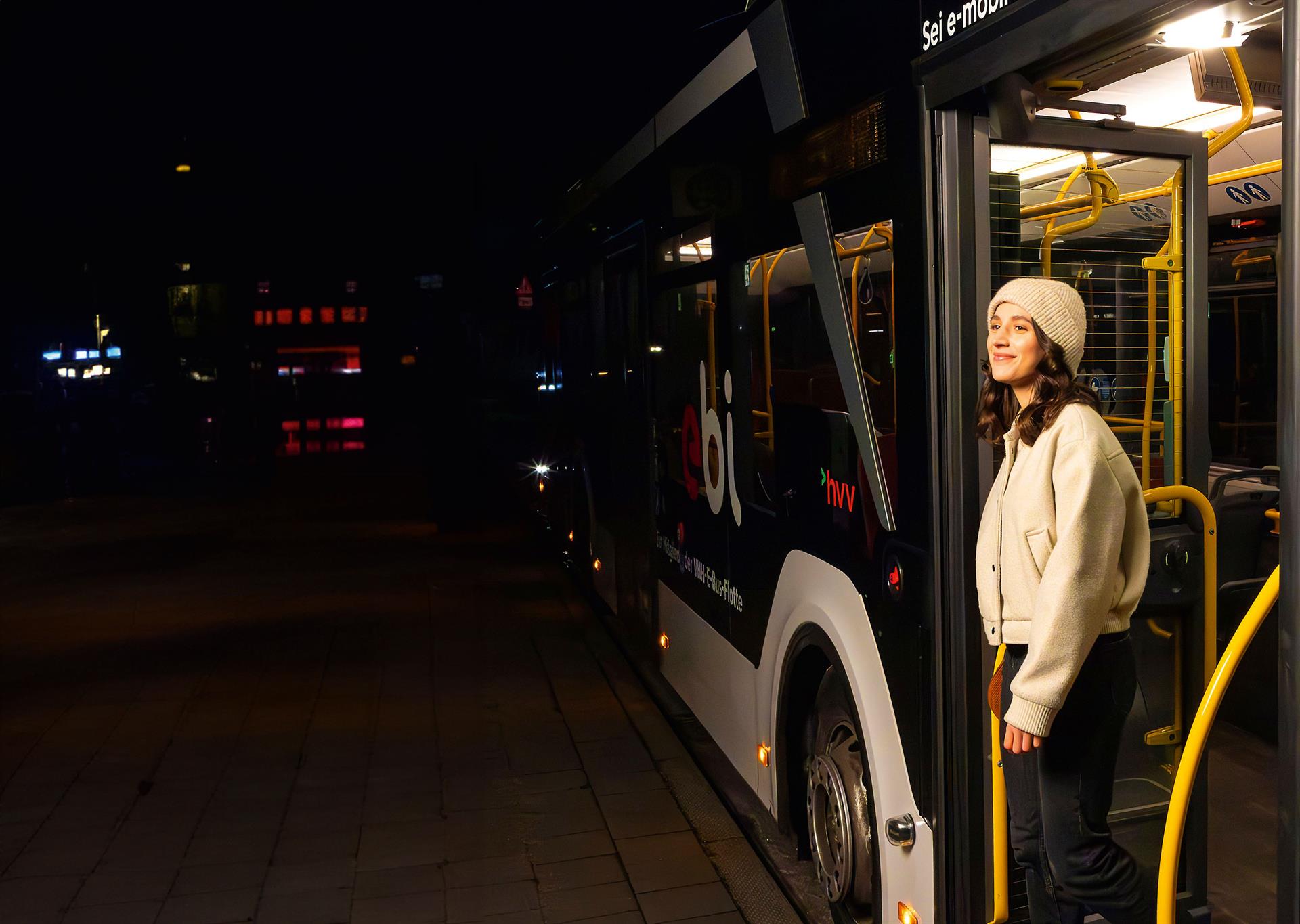 Eine junge Frau mit heller Jacke und Mütze steht nachts in der geöffneten Tür eines hvv-Linienbusses, bereit zum Aussteigen an einem dunklen Straßenrand.