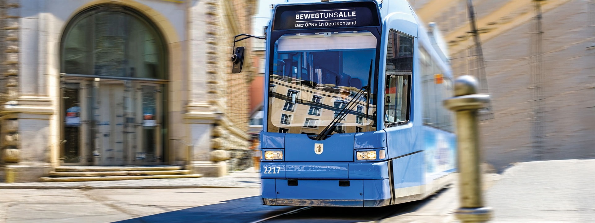 Nahaufnahme einer modernen, hellblauen Straßenbahn in einer sonnigen Innenstadt. Die Bahn fährt in einer leichten Kurve. Auf der Zielanzeige steht der Slogan der VDV-Kampagne „BEWEGT UNS ALLE“ und darunter „Der ÖPNV in Deutschland“.
