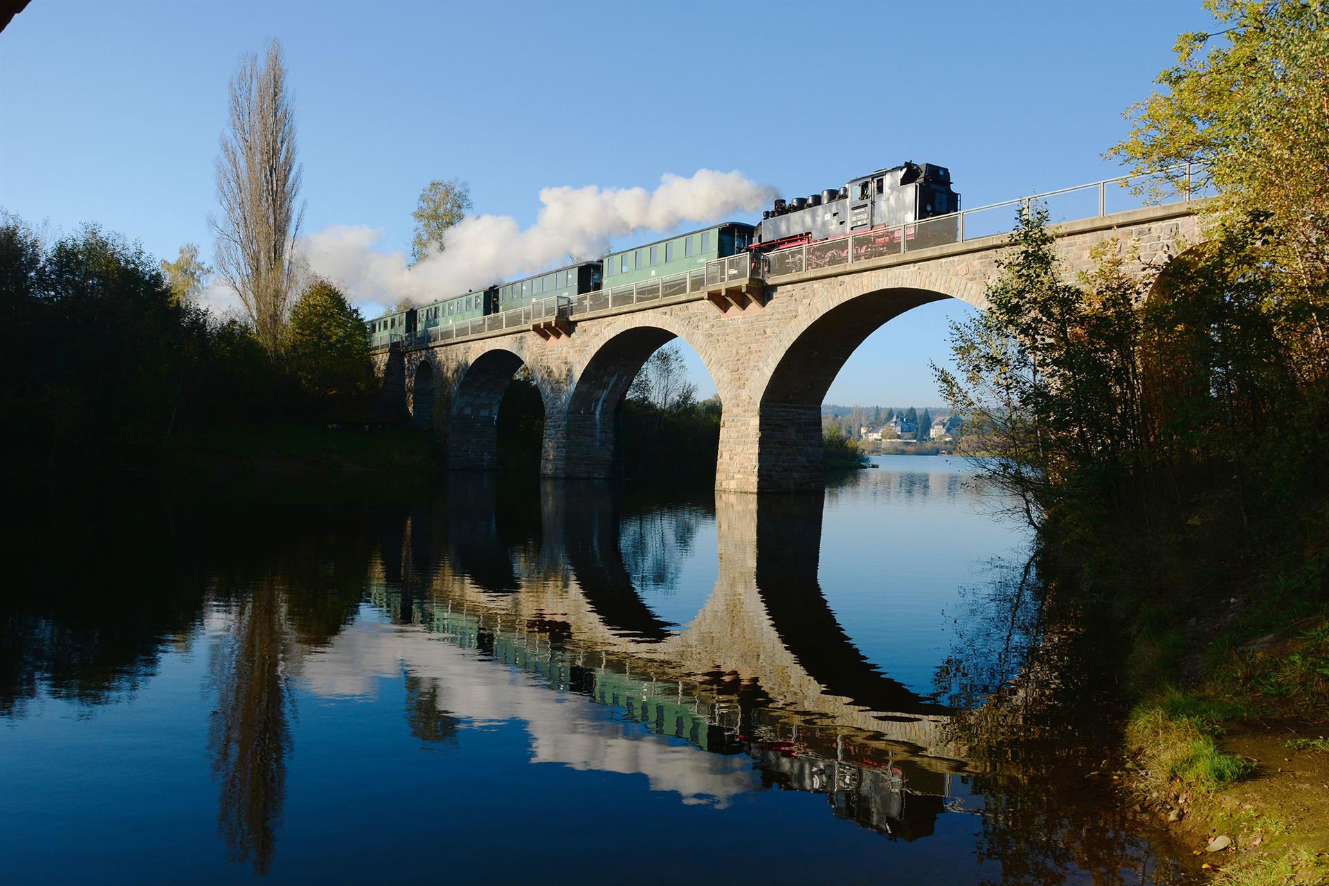 Dampfzug überquert eine mehrbogige Steinbrücke über einem Gewässer, wobei sich Zug und Brücke im Wasser spiegeln.