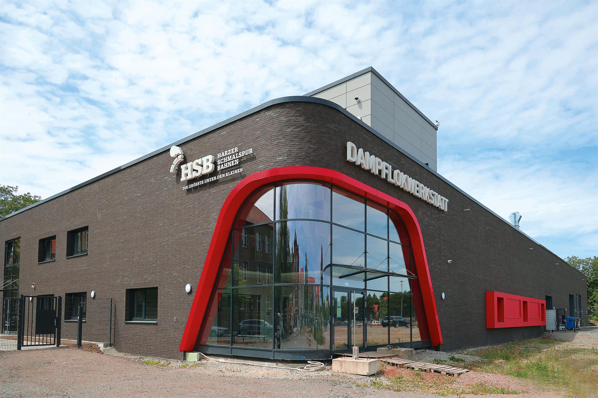 Außenansicht der modernen HSB Dampflokwerkstatt in Wernigerode mit dunkler Klinkerfassade, großem, gerundetem Glasfenster und dem Schriftzug 