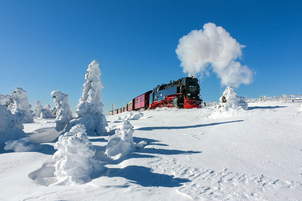 Dampflokomotive 99 7243-1 der Brockenbahn zieht einen Zug durch eine tief verschneite, sonnige Winterlandschaft auf dem Brocken.