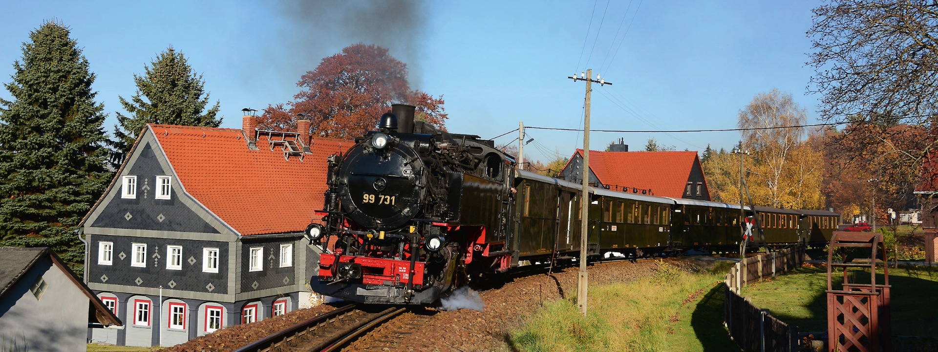 Dampfzug mit Lok 99 731 passiert Wohnhäuser mit roten Dächern in einer Ortschaft der sächsischen Schmalspurbahn.
