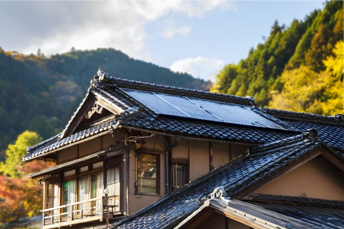 Solaranlage auf dem Ziegeldach eines traditionellen japanischen Holzhauses, im Hintergrund eine sonnige, bewaldete Berglandschaft im Herbst.